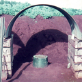 Two toilets dug into red soil divided by wall of wooden boxes and covered by curved corrugated iron panels