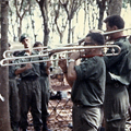 28 October 1969: The band plays for New Zealand and Australian troops amongst a rubber plantation at Fire Support Base Discovery. Men in military uniforms holding musical instruments standing next to tents surrounded by trees
