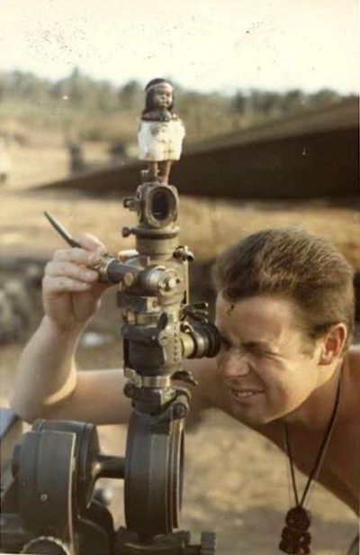 Shirtless man looking through gun sight of artillery gun. A small Māori doll sits above him.