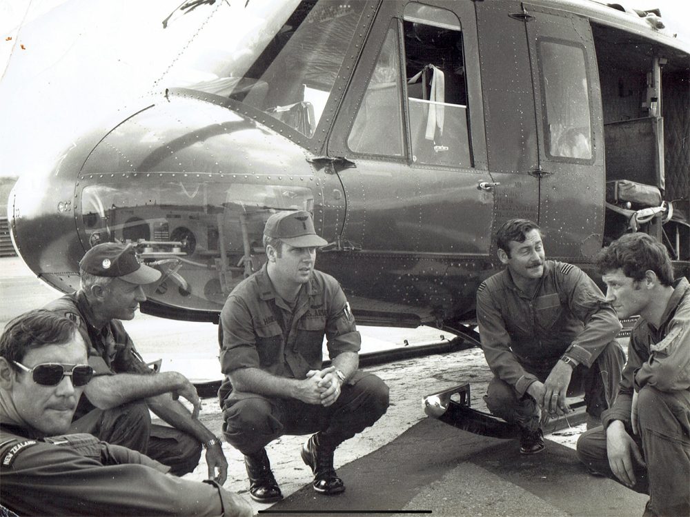 Five men in different military uniforms crouching down in a semi-circle in front of a helicopter