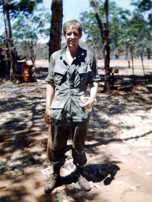 Young man standing in green military uniform in the shade of some trees