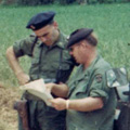 Malcolm Knox Colour photograph of twp men in uniform standing next to jeep vehicle on dirt road.