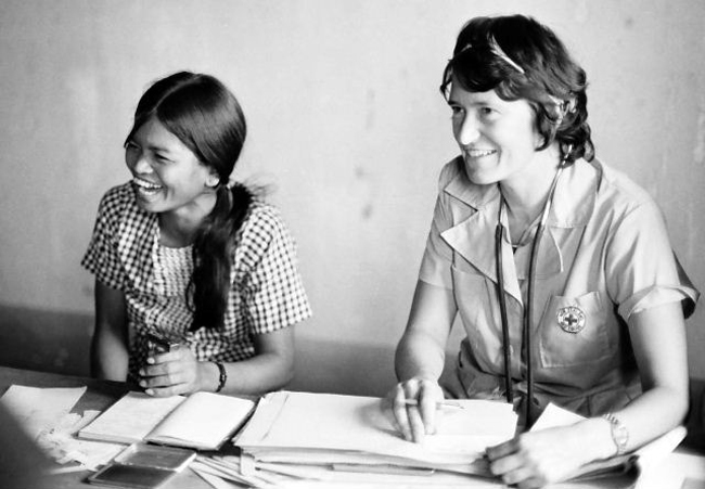 Two women, one wearing a medical uniform and stethoscope, sitting at a desk with paperwork in front of them.