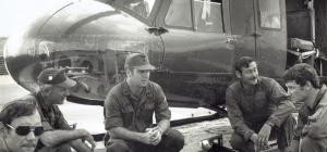 Five men in different military uniforms crouching down in a semi-circle in front of a helicopter