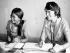 Two women, one wearing a medical uniform and stethoscope, sitting at a desk with paperwork in front of them.