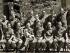 Black and white photograph of men in uniform sitting in two rows. One is holding a white dog.