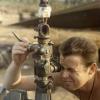 Shirtless man looking through gun sight of artillery gun. A small Māori doll sits above him.