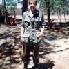 Young man standing in green military uniform in the shade of some trees