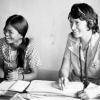 Two women, one wearing a medical uniform and stethoscope, sitting at a desk with paperwork in front of them.