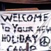 Sign on white sheet saying, 'Welcome to your new holiday camp' next to a peace sign