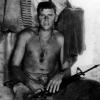 Shirtless soldier sitting in tent with gun on his lap