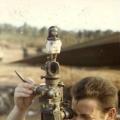 Shirtless man looking through gun sight of artillery gun. A small Māori doll sits above him.