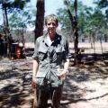 Young man standing in green military uniform in the shade of some trees