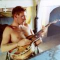 Bare chested man sitting at table holding a guitar.