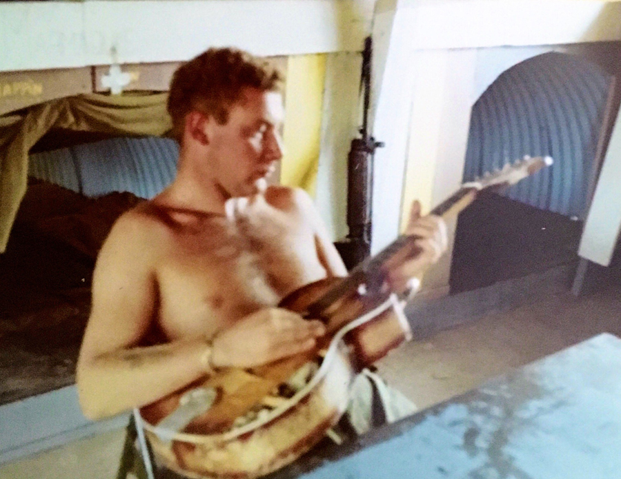 Bare chested man sitting at table holding a guitar.